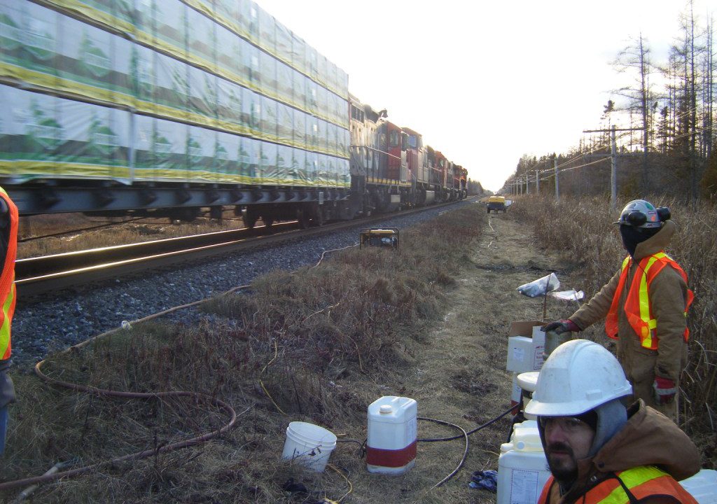 Grouting crew stabilizing a rail embankment beside a passing freight train on an active line